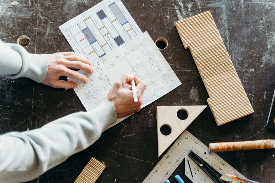 Hände entwerfen in einer Werkstatt mit Holzwerkzeugen architektonische Entwürfe auf Papier.